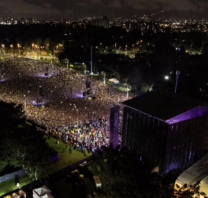 Aerial view of a large outdoor concert at night, capturing the vibrant energy typical of LATAM music festivals, with a dense crowd in front of a brightly lit stage surrounded by trees and city lights in the background.